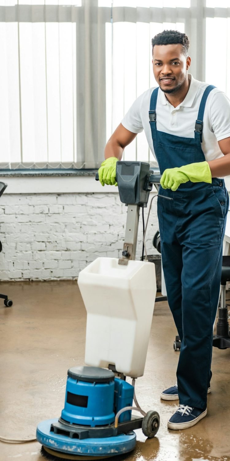 smiling african amercan cleaner looking at camera while washing floor with cleaning machine